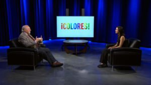 Two people sit in armchairs on a studio set with a screen displaying the word “¡COLORES!” in colorful letters.