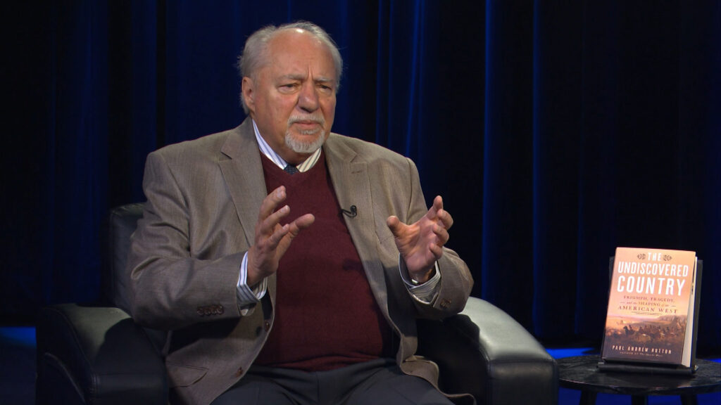 An older man in a suit sits in a black armchair gesturing with his hands; a book titled "The Undiscovered Country" is displayed on a small table beside him.