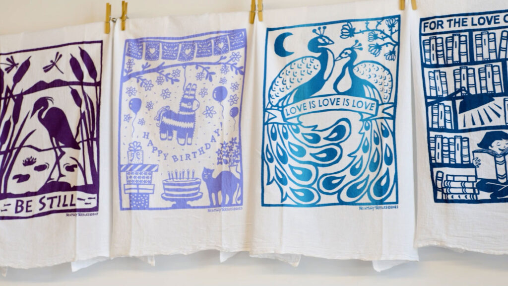 Four printed fabric panels with blue illustrations hang side by side on a line. Designs include a bird, a birthday scene, peacocks with “Love Is Love Is Love,” and a library scene.