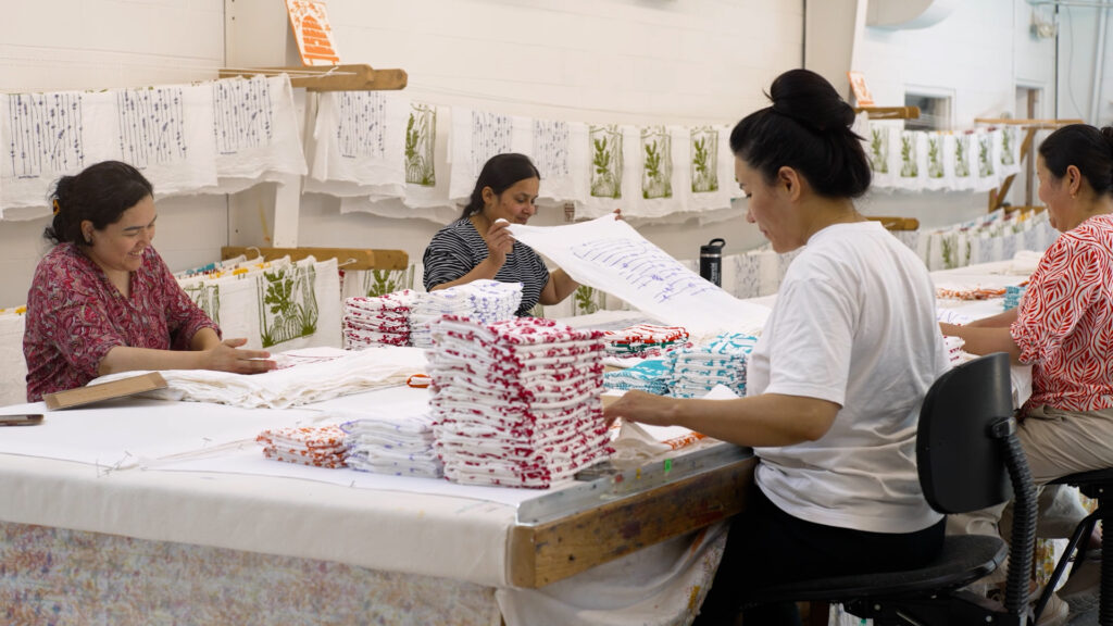 Three women sit at a table folding and sorting printed textiles in a well-lit workspace, with stacks of fabric and hanging cloths in the background.
