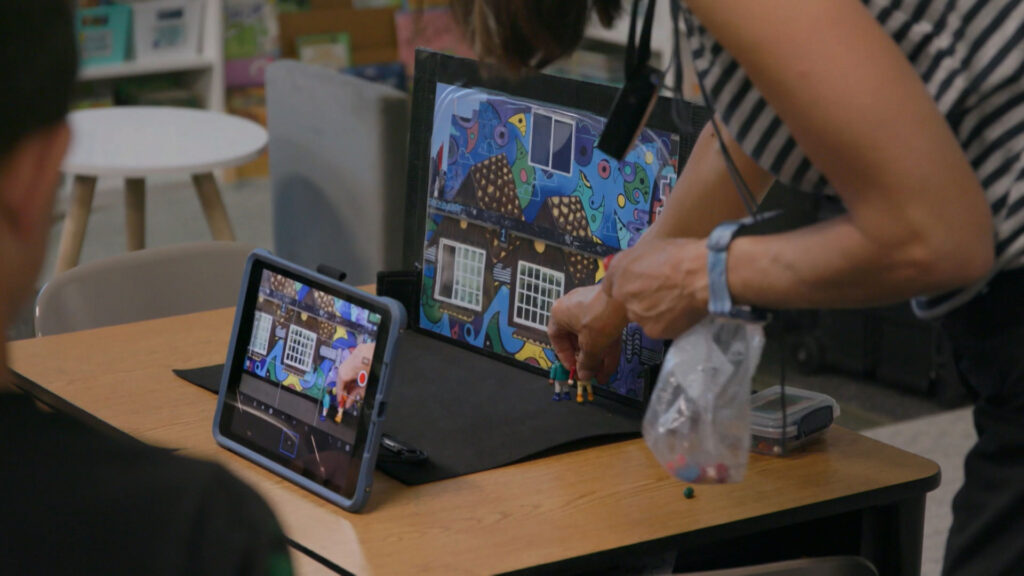 A person arranges small figurines in front of a colorful backdrop while a tablet records the scene on a classroom desk.