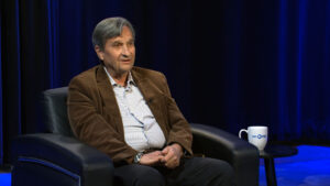 An older man in a brown corduroy jacket sits on a black armchair, speaking, with a white mug on a small table beside him. The background is dark blue with curtains.