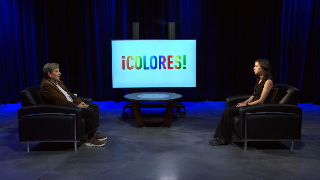 Two people sit facing each other in a studio with a round table between them and a screen displaying the word "¡COLORES!" in colorful letters.