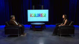 Two people sit facing each other in a studio with a round table between them and a screen displaying the word "¡COLORES!" in colorful letters.