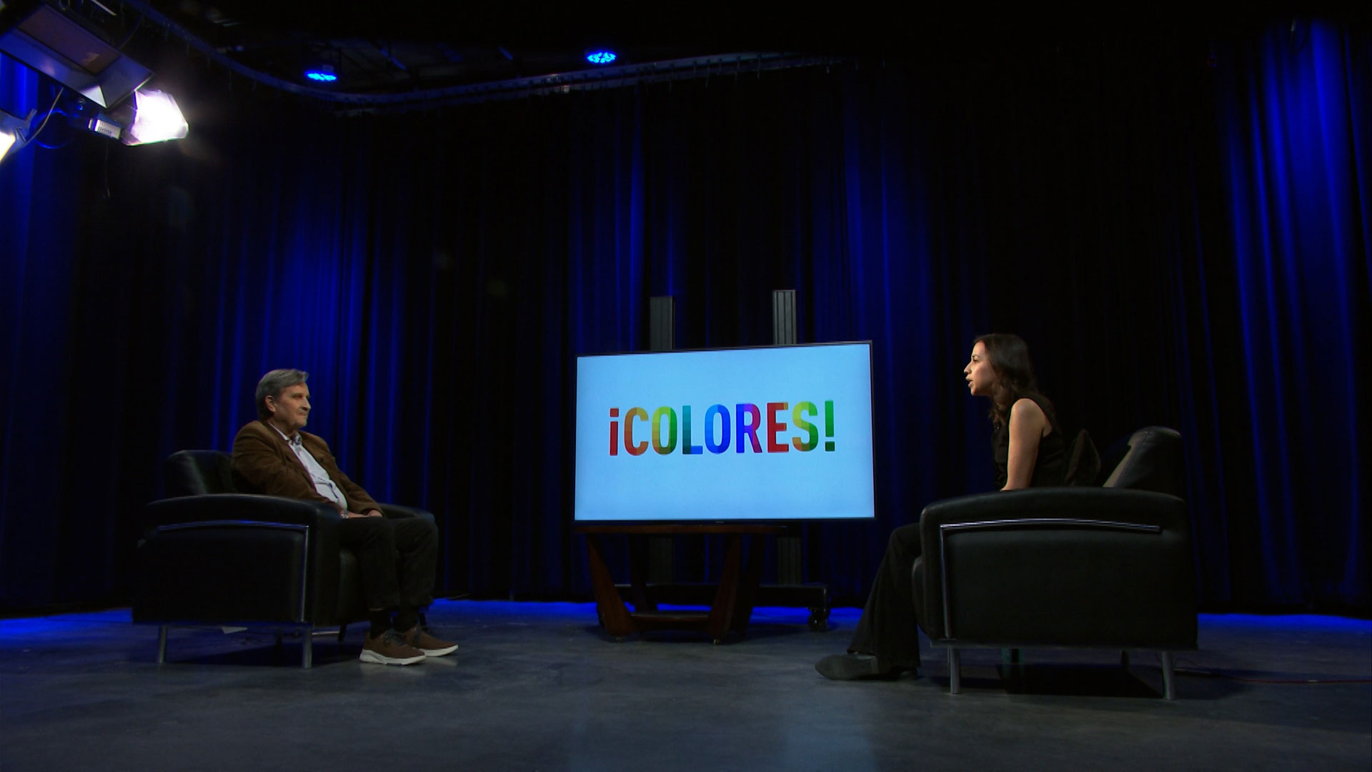 Two people sit across from each other in a dark studio with blue lighting; a screen between them displays the word "¡COLORES!" in colorful letters.