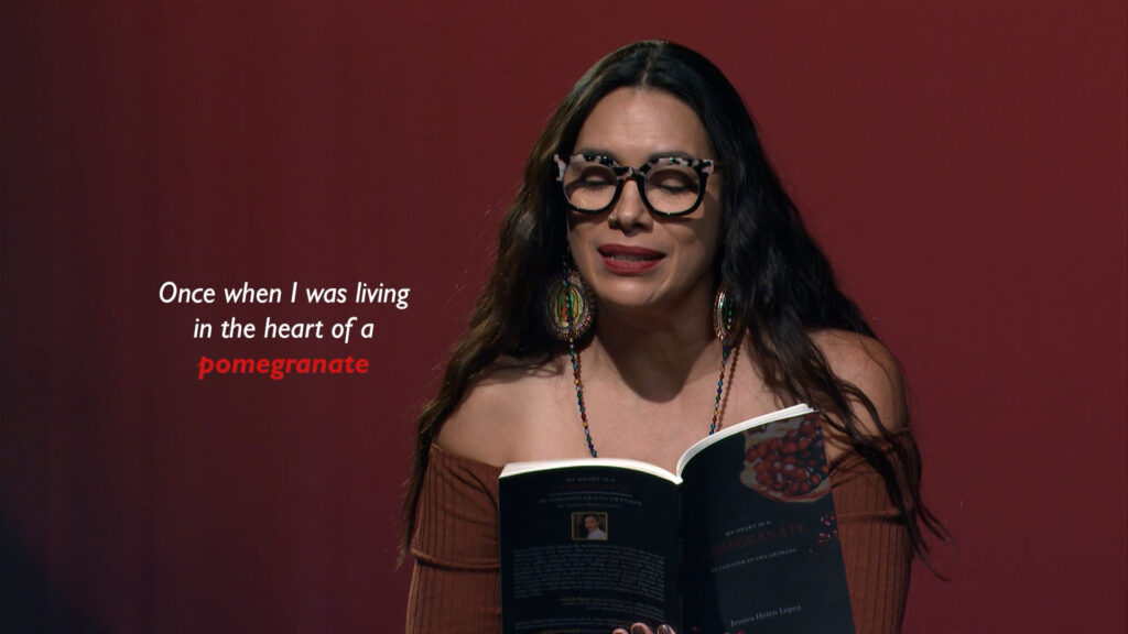 A woman with long dark hair and large glasses reads aloud from a book against a red background with a projected quote about a pomegranate.