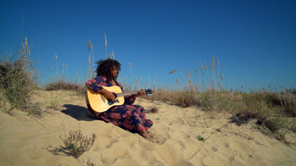 A person in a floral dress sits barefoot on sandy dunes, playing an acoustic guitar, with grass and a clear blue sky in the background.