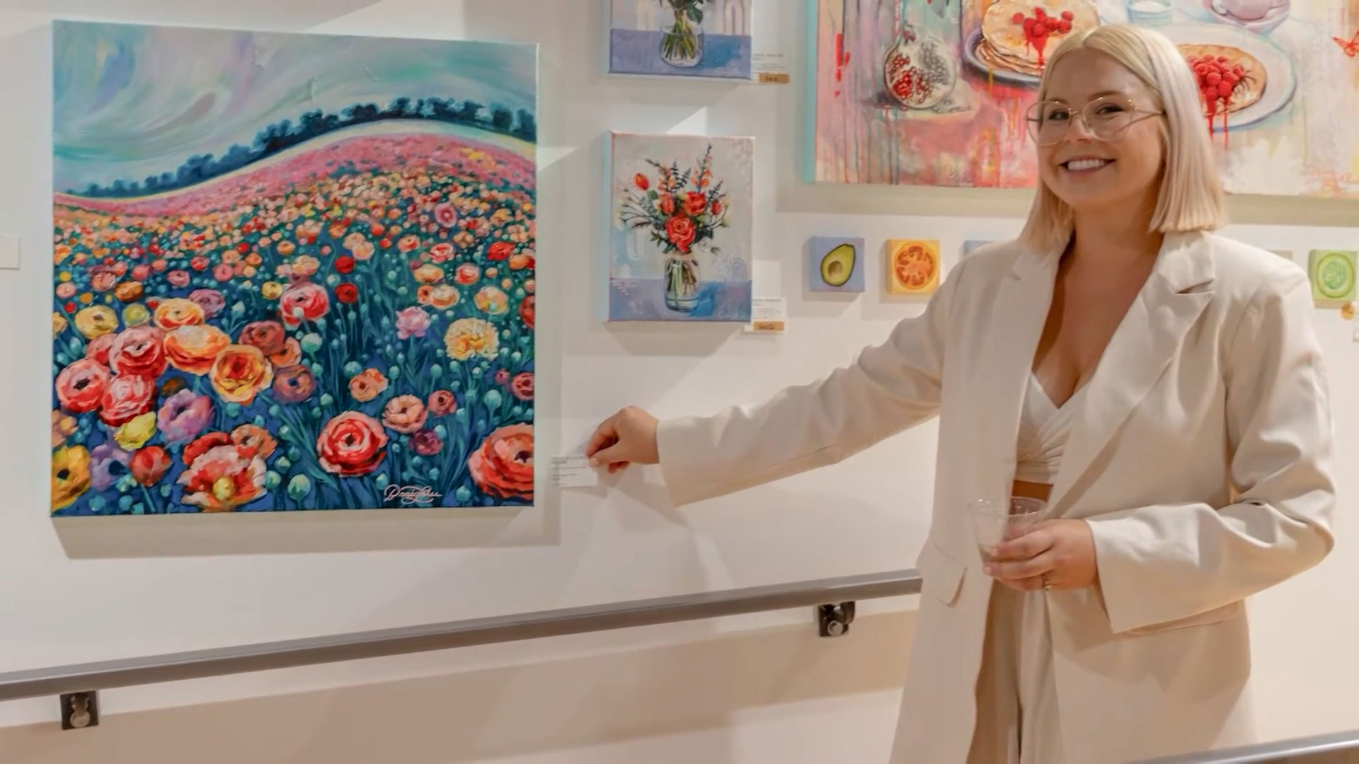 A woman in a light-colored outfit stands indoors, smiling and pointing at a colorful floral painting displayed on a gallery wall with other artworks.