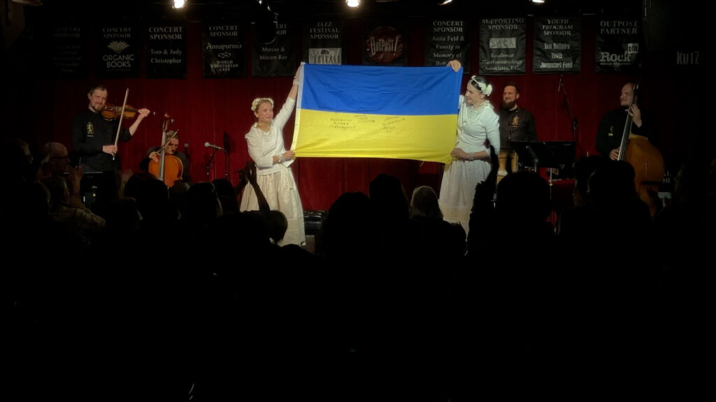 Performers on stage hold up a large Ukrainian flag in front of an audience, with musicians playing string instruments in the background.