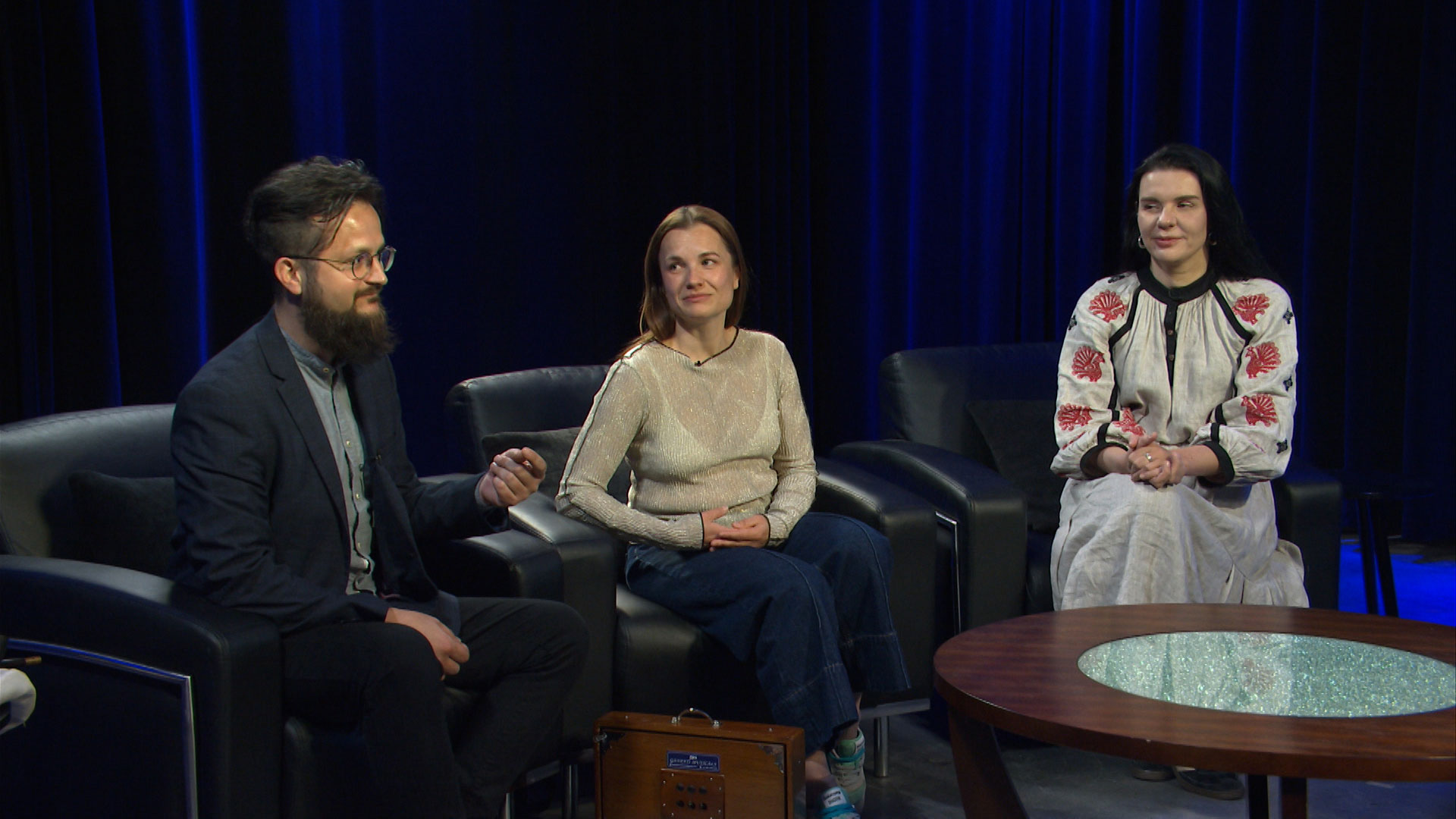 Three people sit on black armchairs in a studio with dark curtains and blue lighting, engaged in conversation near a round table.