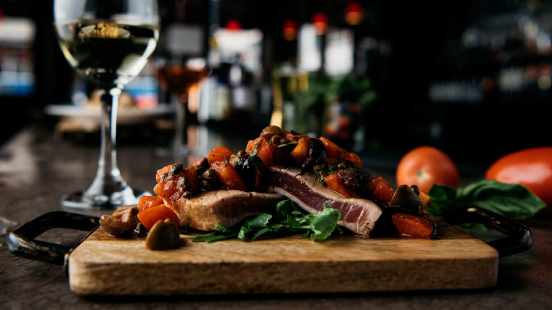 A seared tuna steak topped with tomato and olive garnish sits on a wooden board with greens, next to a glass of white wine in a dimly lit restaurant setting.