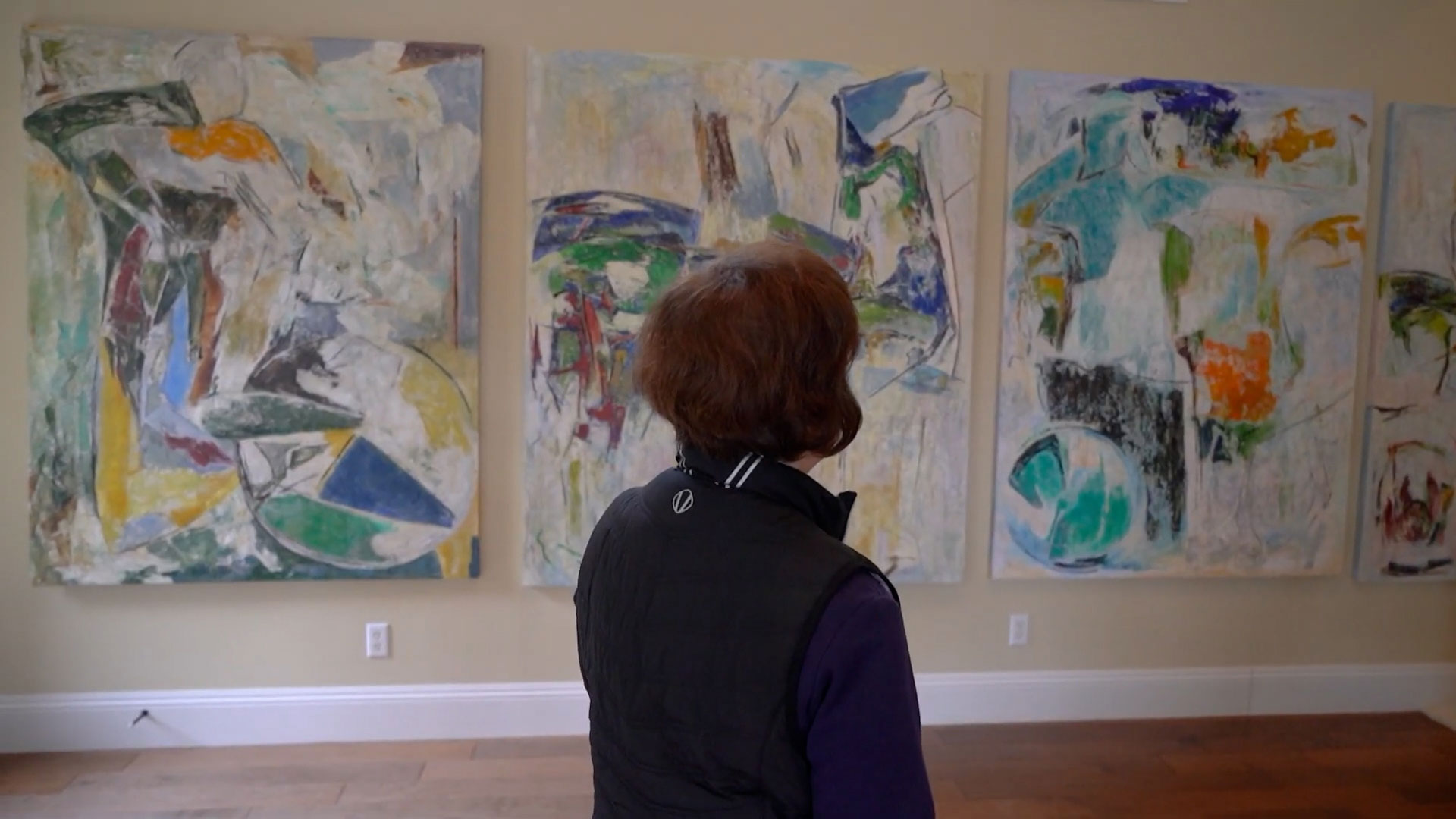 A person stands indoors, facing three large abstract paintings with blue, green, and yellow tones displayed on a beige wall.