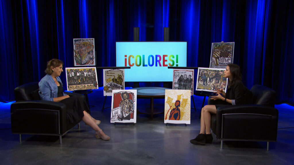 Two people sit in armchairs on a studio set with artwork displayed between them and a screen behind them reading "¡COLORES!" in colorful letters.
