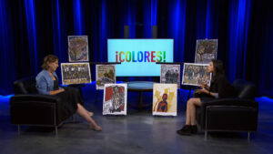 Two people sit in armchairs on a studio set with artwork displayed between them and a screen behind them reading "¡COLORES!" in colorful letters.