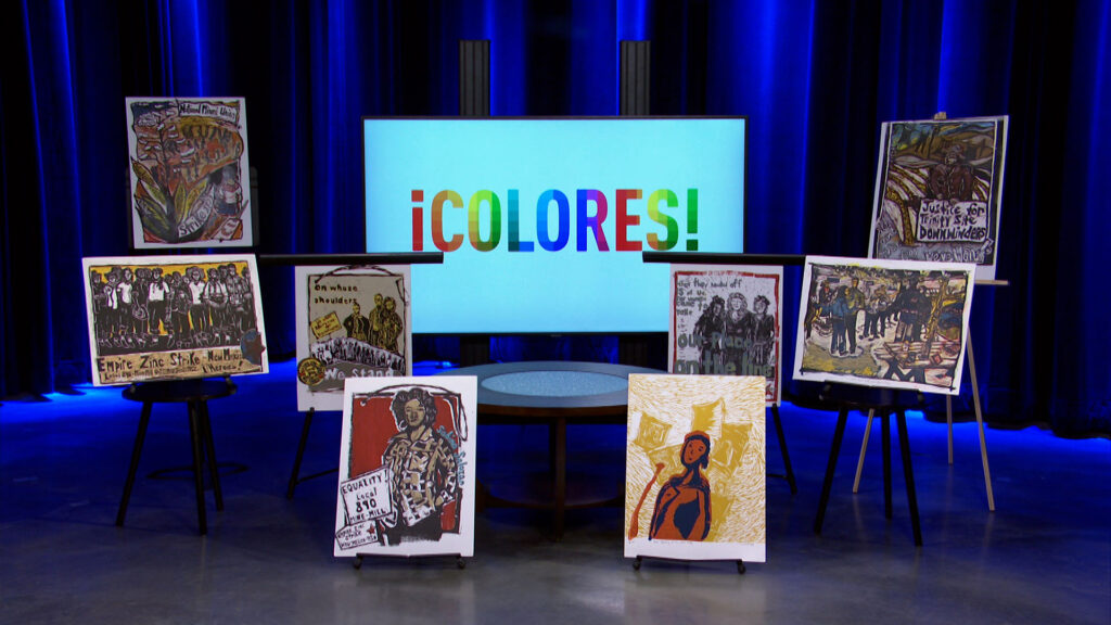 Several colorful artworks on easels are arranged on a stage with a large screen in the background displaying the text “¡COLORES!” in multicolored letters.