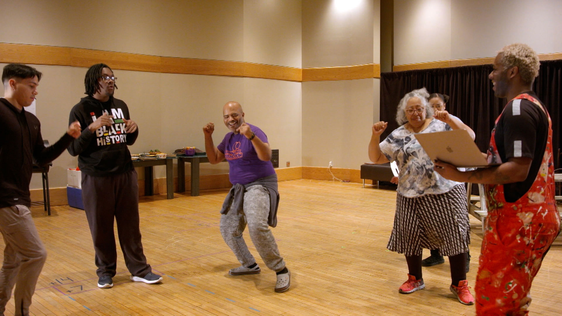 Five people stand in a room with wooden floors, laughing and posing enthusiastically, while one person holds a laptop. A table with items is visible in the background.