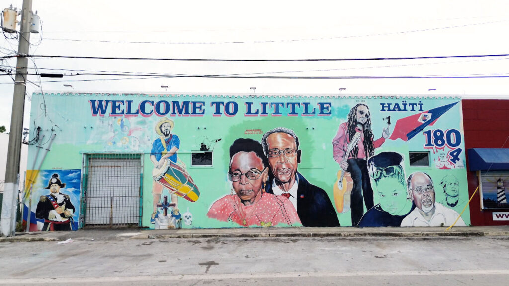 A colorful mural on a building wall reads "Welcome to Little Haiti" and features portraits of notable Haitian figures and symbols.