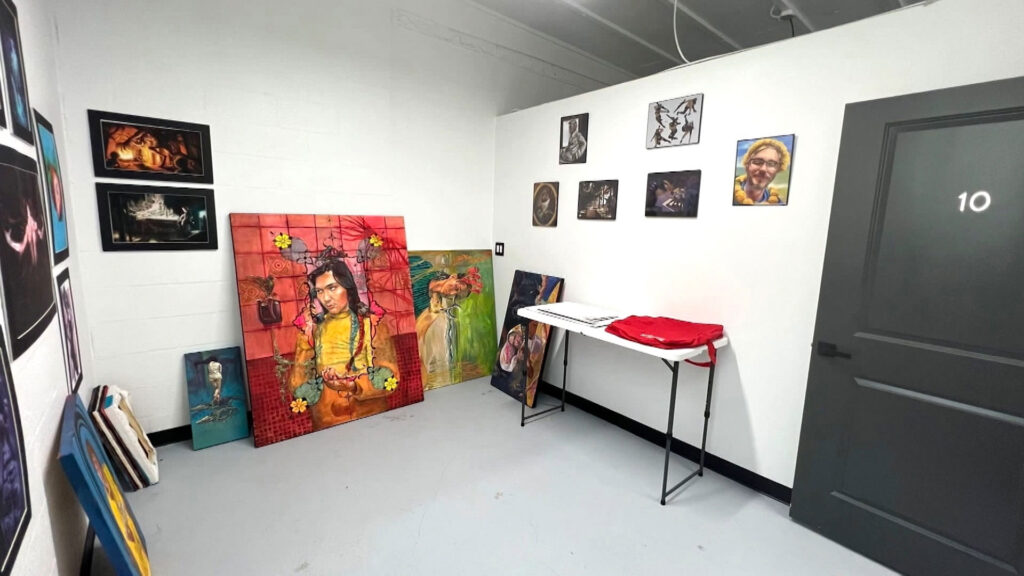 A small art studio with paintings and portraits on white walls, several canvases leaning against the wall, a folding table, and a closed gray door labeled “10.”.