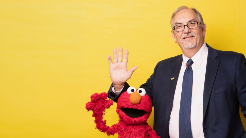 A man in a suit and tie stands next to the Elmo puppet, both waving, in front of a plain yellow background.