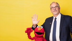 A man in a suit and tie stands next to the Elmo puppet, both waving, in front of a plain yellow background.