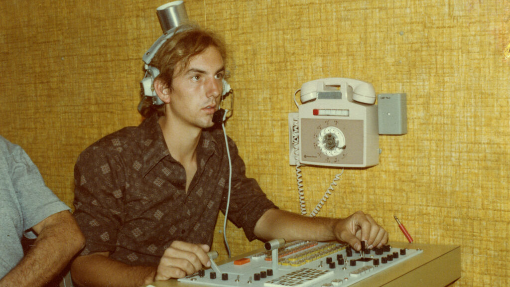 Young man operating audio equipment with a headset, sitting at a control panel; a rotary wall phone is mounted above the desk against a textured yellow wall.