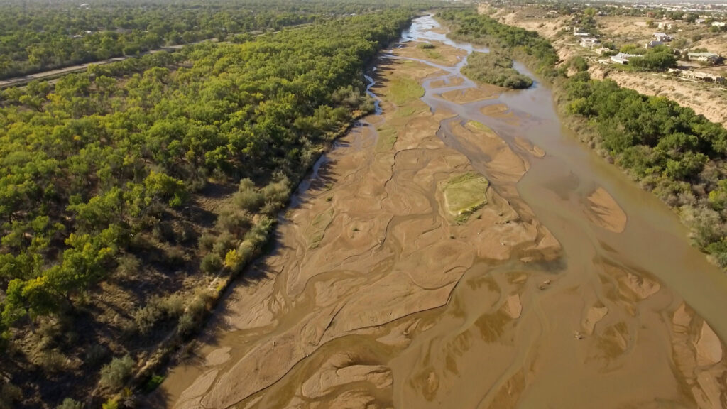 Aerial view of a mostly dry riverbed with patches of water, bordered by dense green trees on one side and sparse vegetation on the other.
