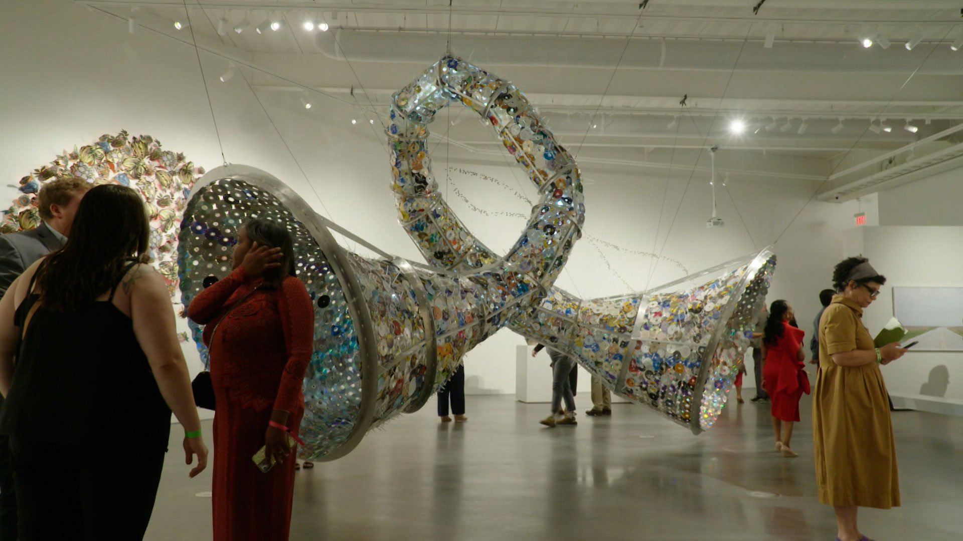 People view a large, suspended sculptural installation made of recycled materials inside a modern art gallery.