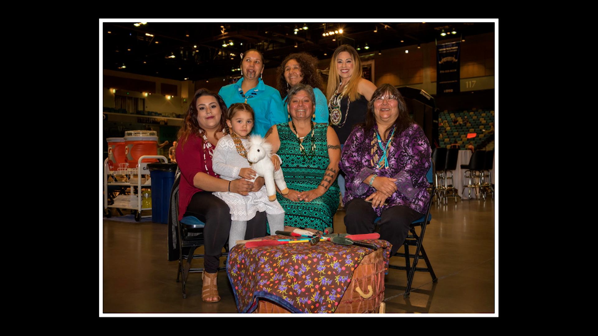 Seven women, including a young girl holding a stuffed animal, pose together indoors at an event, seated and standing around a table with a patterned cloth.