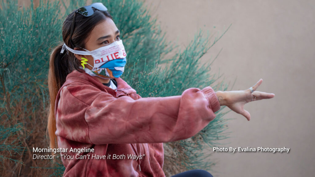 A woman in a pink jacket and colorful face mask gestures with her hand while seated outdoors near green bushes. Text on image credits Morningstar Angeline and photographer Evalina.