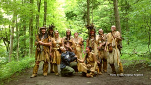 Nine people, most wearing tan leather clothing and nature-inspired accessories, pose and smile together in a green forest. Text in the corner credits the photo to Morningstar Angeline.