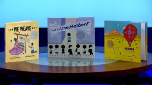 Three children's books are displayed upright on a round table: "We Weave," "Fall in Line, Holden!," and "Herizon," each featuring an award medal on the cover.