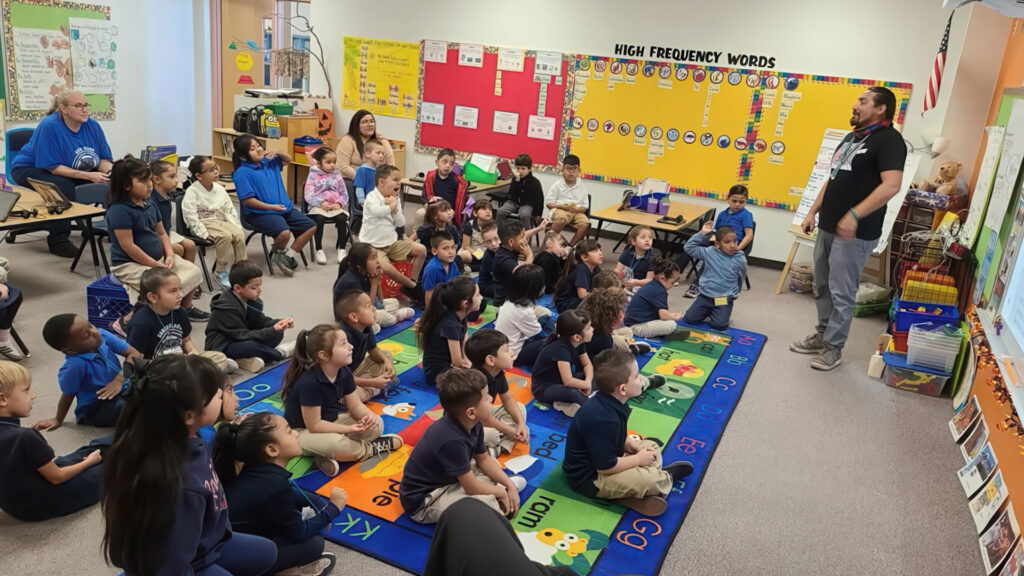 A teacher stands at the front of a classroom while a group of young students sit on a colorful alphabet rug, attentively listening. Educational posters and high frequency words cover the walls.