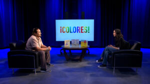 Two people sit across from each other in a studio with blue lighting. A table with books and a screen displaying "¡COLORES!" are between them.