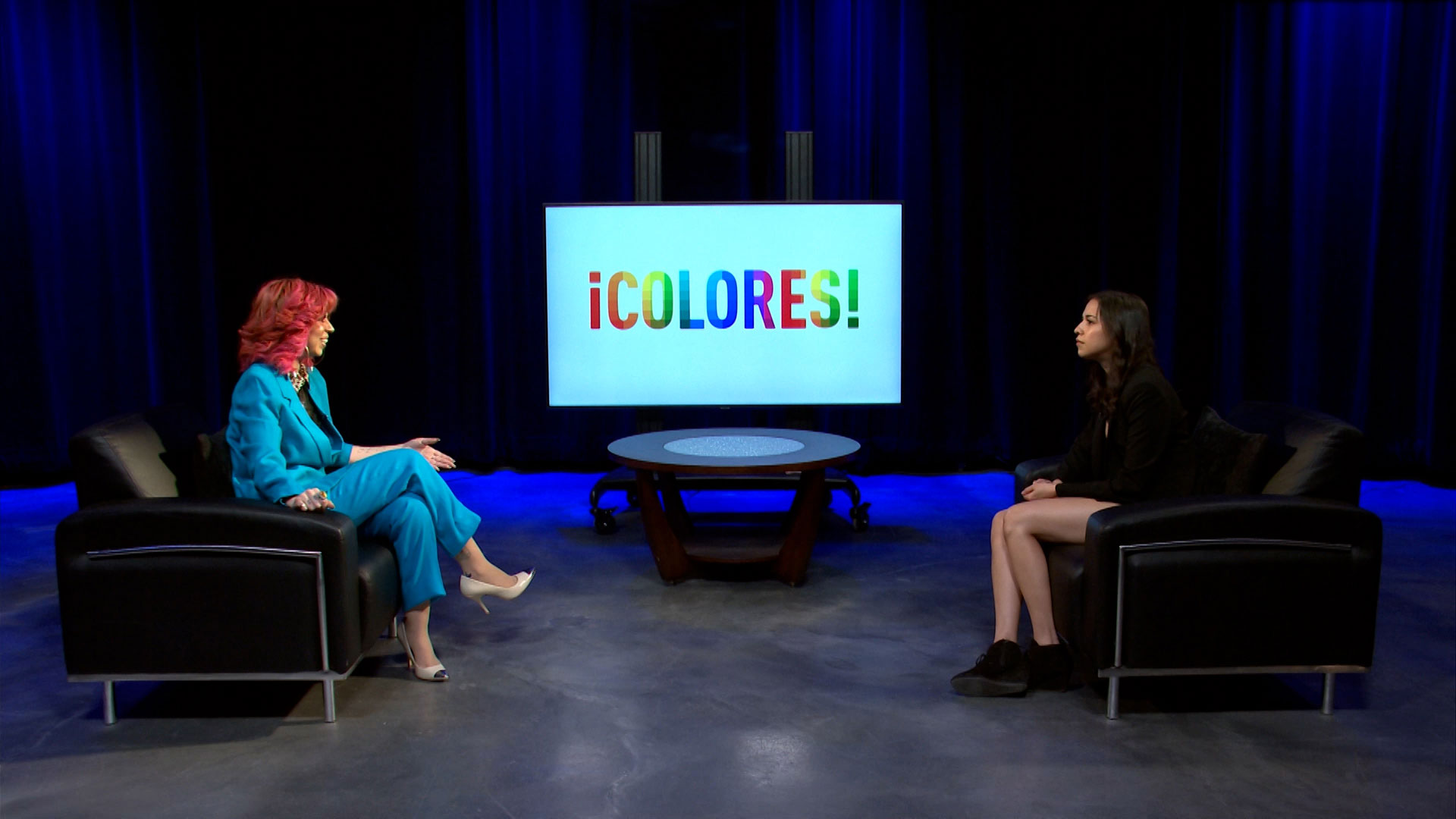 Two people sit across from each other in a studio with a screen behind them displaying the word "¡COLORES!" in colorful letters.