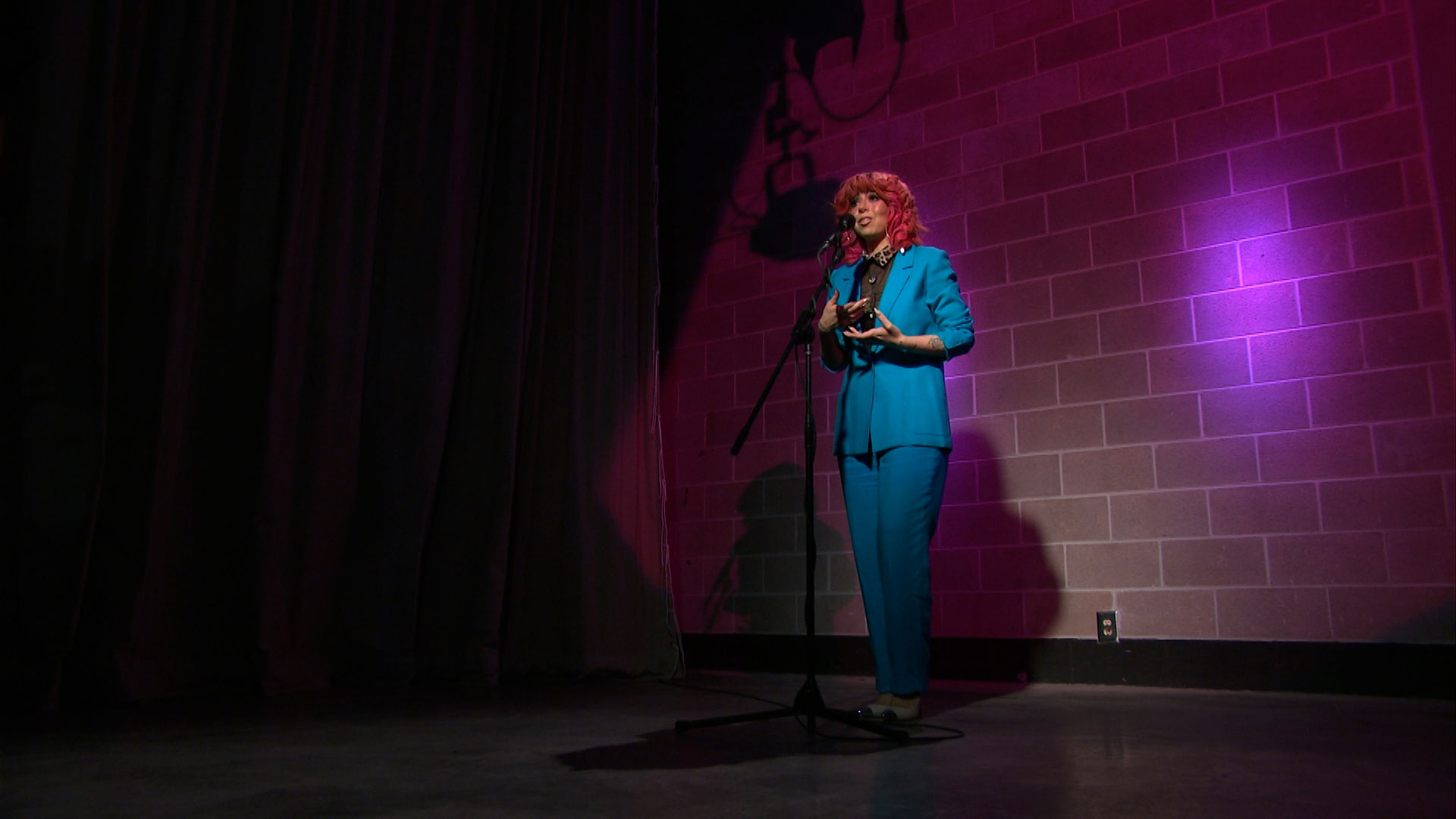 Person in a bright blue suit stands on stage under pink lighting, speaking or performing into a microphone. Brick wall and curtains are visible in the background.