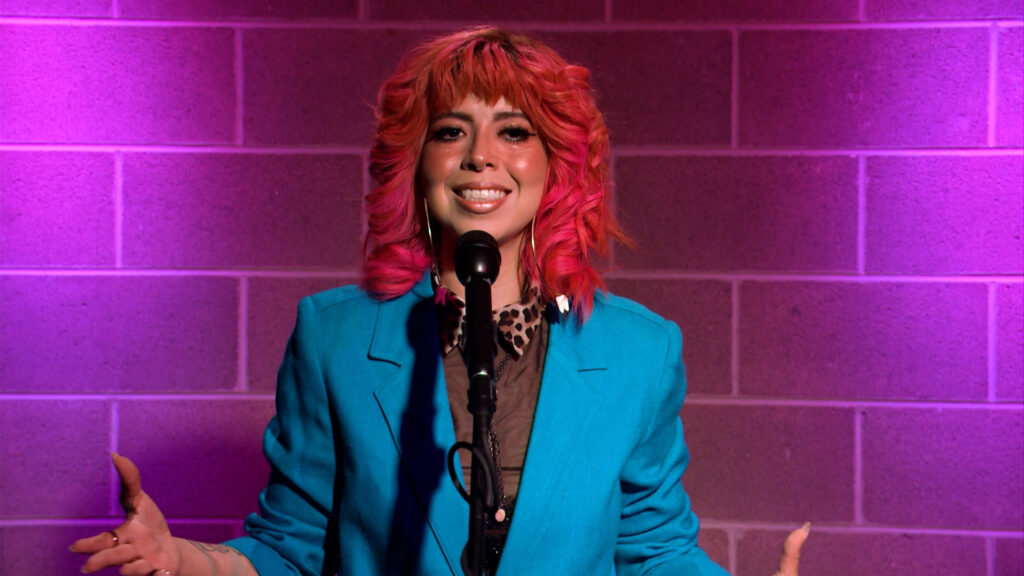 A person with bright pink hair wearing a blue blazer speaks into a microphone in front of a brick wall with purple lighting.