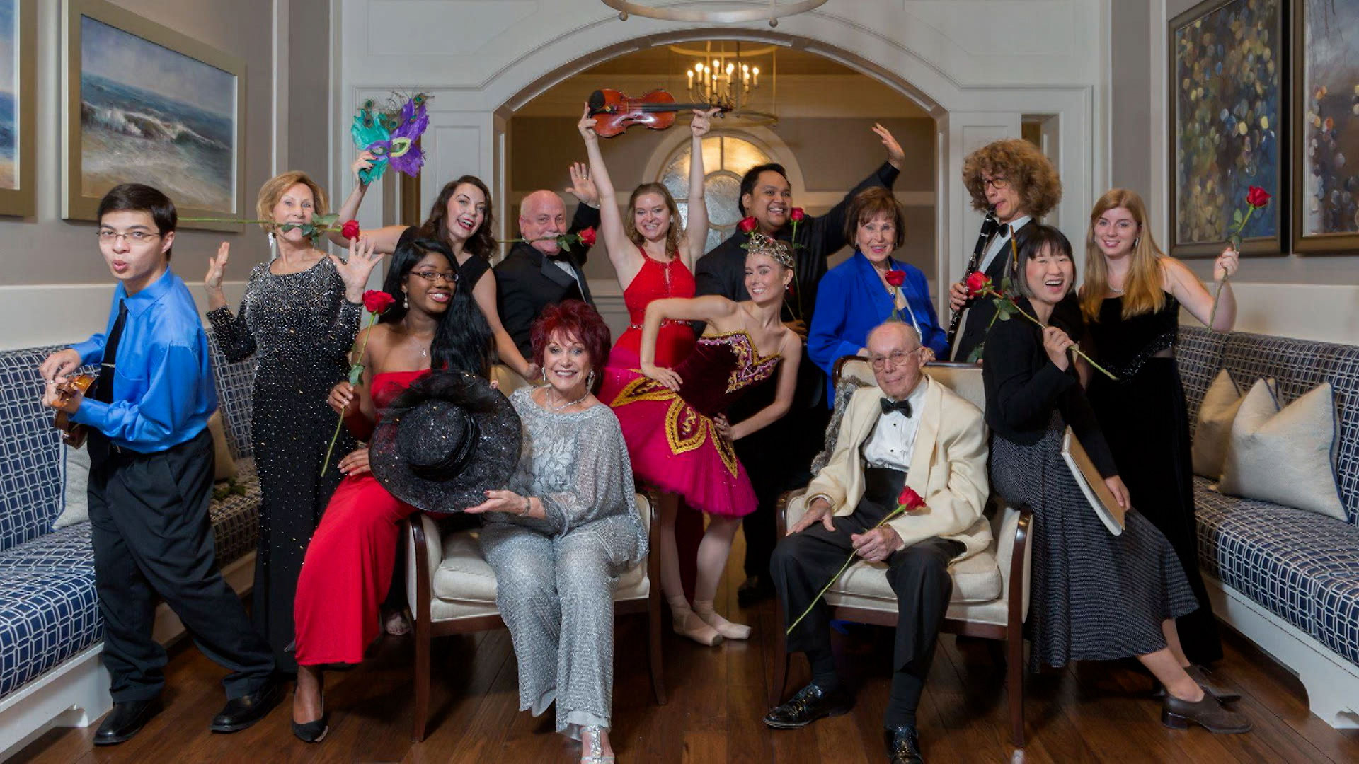 A diverse group of people dressed in formal and performance attire pose indoors, some holding instruments, masks, and roses, with cheerful and expressive gestures.