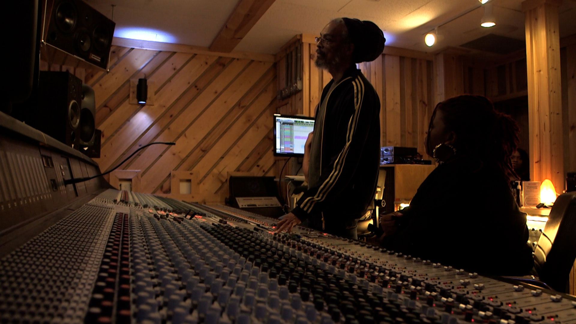 Two people are working at a large audio mixing console in a dimly lit recording studio with wooden panel walls and computer monitors in the background.