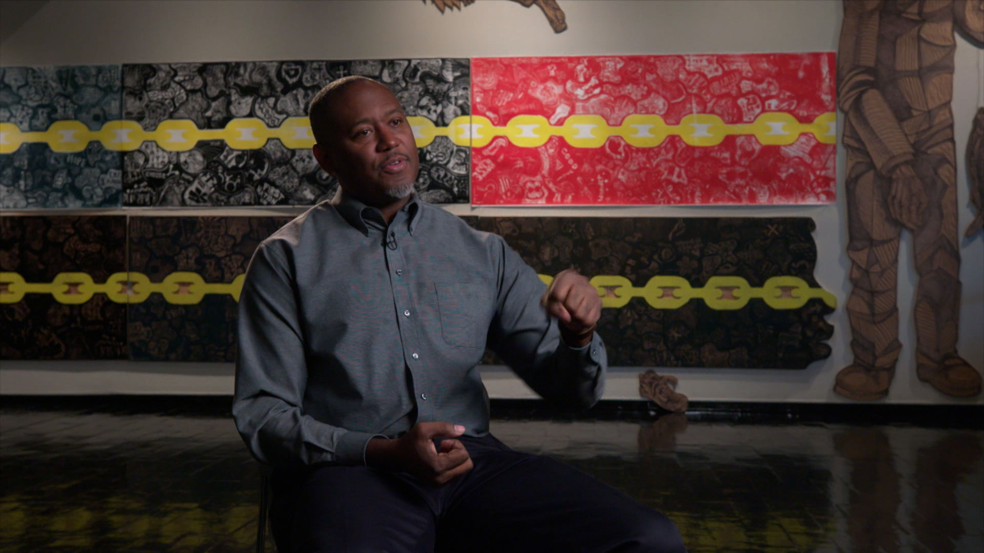 A man sits and gestures while speaking in front of a wall displaying colorful, patterned artwork with yellow chain designs.