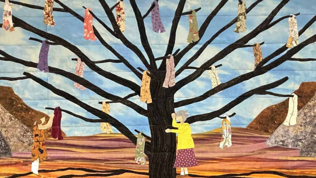 A fabric artwork shows two women near a large tree with colorful dresses hanging from its branches, set against a landscape with mountains and a blue sky.