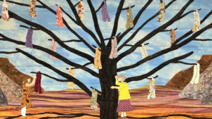 A fabric artwork shows two women near a large tree with colorful dresses hanging from its branches, set against a landscape with mountains and a blue sky.