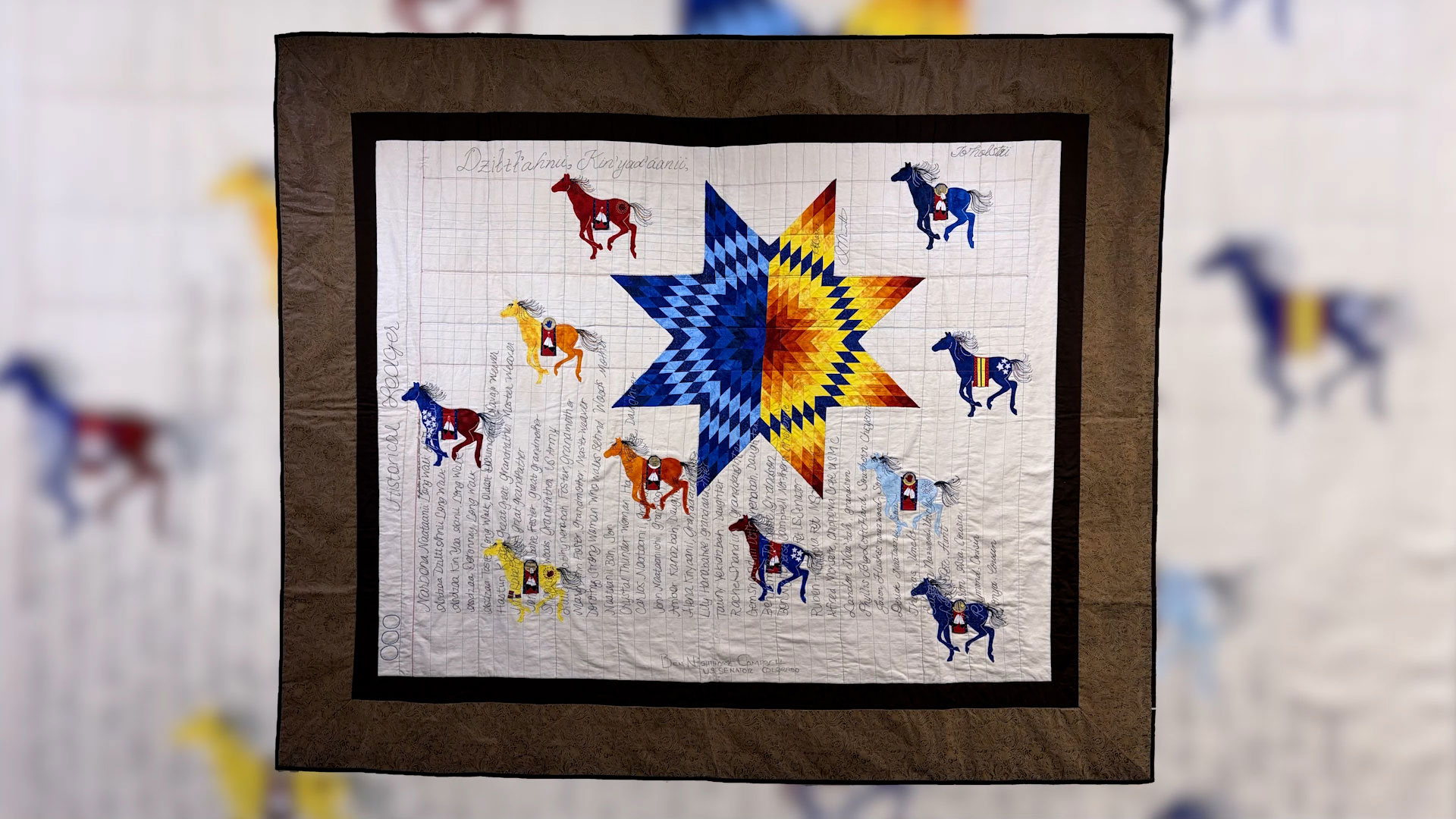 A quilt featuring a large, multicolored star in the center, surrounded by twelve embroidered horses and bordered with brown and black fabric.
