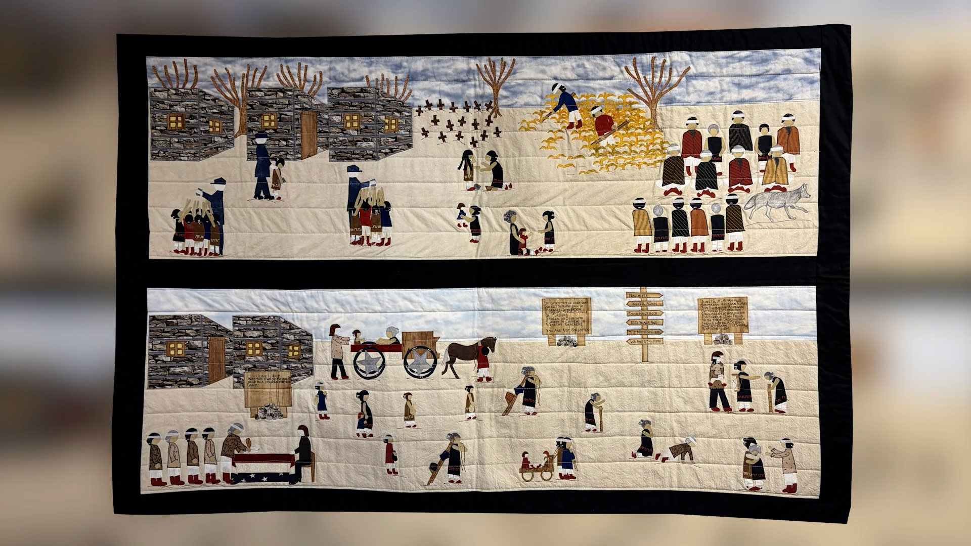 A handmade quilt featuring two panels with stitched scenes of people in historical clothing performing daily activities in village settings, with buildings, animals, trees, and carts.