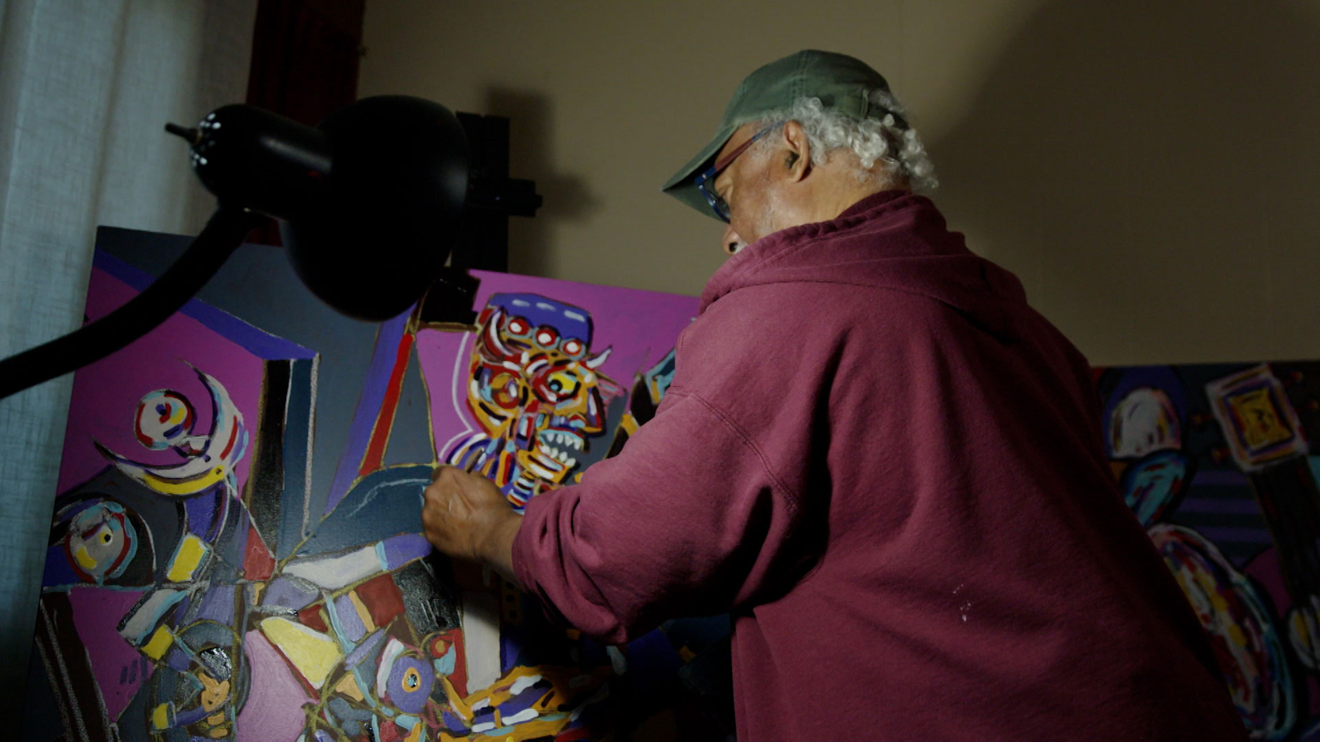 An older man wearing a cap and maroon hoodie paints a colorful, abstract artwork on a canvas under a lamp.