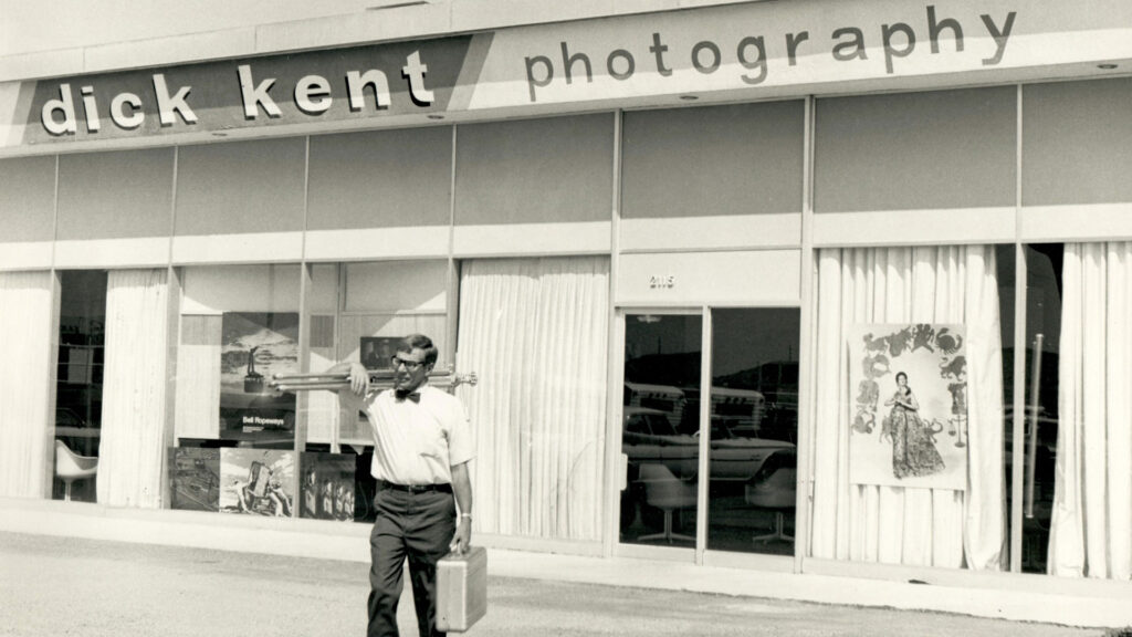 A man carrying a suitcase and equipment walks past the storefront of "dick kent photography," which has large windows displaying photos and a poster.