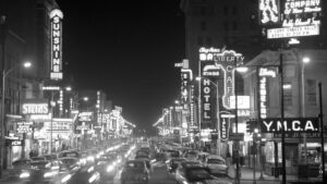 Black and white photo of a busy city street at night with heavy traffic and numerous illuminated neon signs for hotels, restaurants, theaters, and businesses.