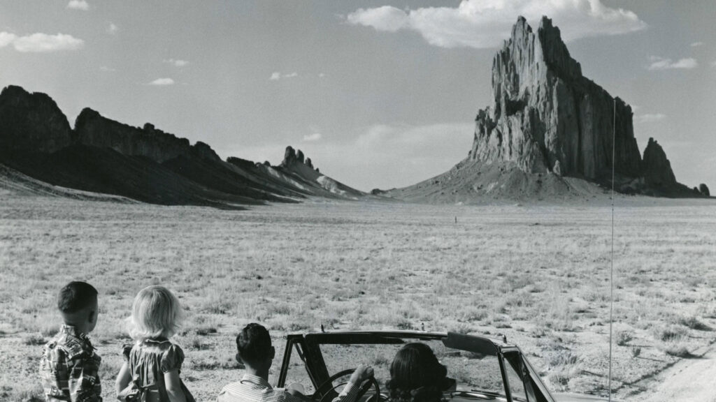 Four children in a convertible face a large rock formation in a flat, arid landscape under a partly cloudy sky.