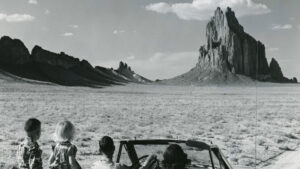 Four children in a convertible face a large rock formation in a flat, arid landscape under a partly cloudy sky.