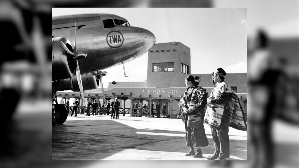 Two people in traditional clothing stand near a TWA airplane at an airport terminal labeled "Albuquerque," with other people in the background.