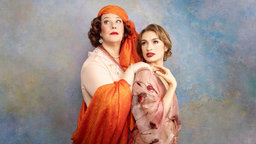 Two women in vintage-style clothing, one in an orange scarf and the other in a light pink dress, pose against a textured blue background, both looking slightly to the side.